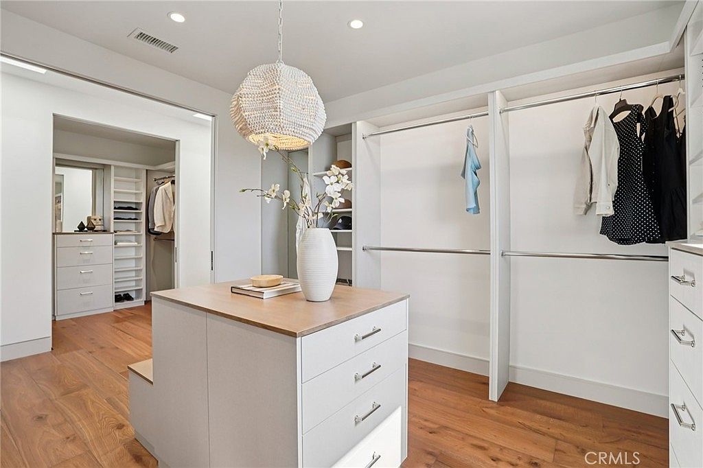 This spacious walk-in closet features a central island with a light wood top and white drawers, providing ample storage and a sophisticated focal point. The room is equipped with extensive built-in shelving and hanging rods, illuminated by a decorative textured pendant light. The warm wood flooring and neutral color palette create an inviting and organized atmosphere, with a secondary closet area visible through the doorway in the background.