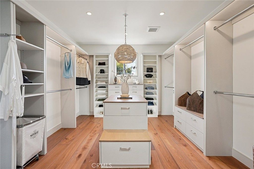 This spacious, well-organized walk-in closet features custom white cabinetry with a mix of hanging rods, open shelving, and drawers. A central island provides additional storage and a surface for accessories, topped by a decorative vase, while a textured pendant light hangs from the ceiling. The room is finished with warm-toned wood flooring, creating a bright and inviting atmosphere.