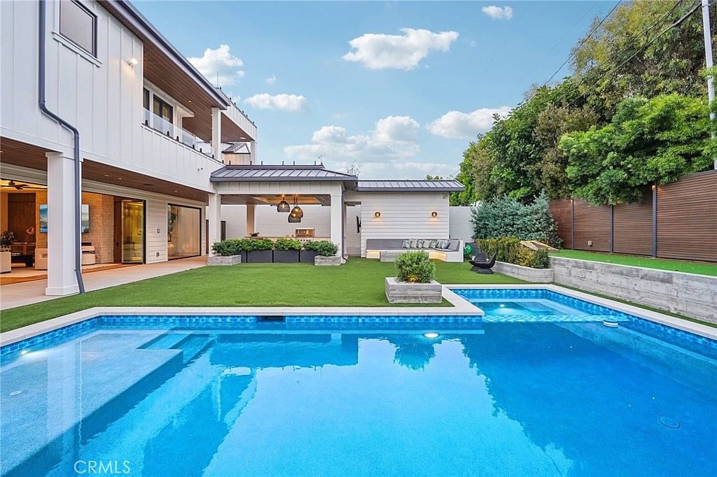 This bright, modern backyard features a pristine swimming pool with an integrated spa, set against a backdrop of a contemporary white home. A manicured artificial turf lawn separates the pool area from a covered outdoor kitchen and lounge space, which includes built-in seating and stylish pendant lighting. The scene is framed by a tall wooden privacy fence and lush greenery, creating a serene and luxurious outdoor living environment.