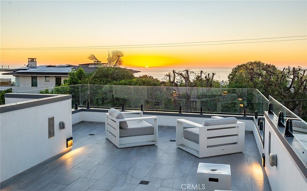 This modern rooftop deck offers a panoramic sunset view over the ocean, framed by a sleek glass railing. The space is furnished with two white lounge chairs and a small side table, set against a backdrop of lush trees and a neighboring residence. The clean, minimalist aesthetic and elevated perspective create a serene and luxurious outdoor living environment.