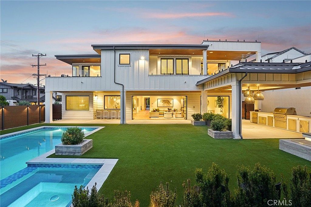 This modern luxury residence features a clean, white-paneled exterior with expansive glass doors that open to a seamless indoor-outdoor living space. The backyard includes a pristine swimming pool with an integrated spa, a manicured artificial turf lawn, and a covered outdoor kitchen area equipped with stainless steel appliances. The scene is captured at dusk, highlighting the warm interior lighting and the sophisticated, contemporary architectural design.