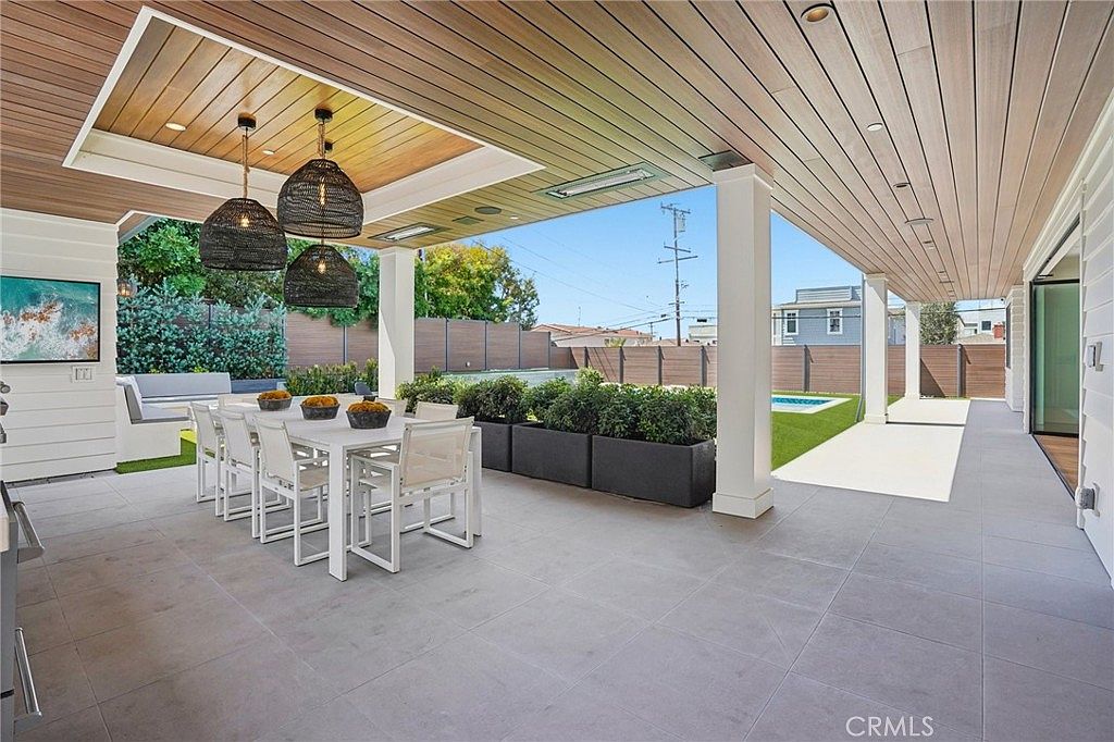 This expansive covered patio features a sleek, modern design with large-format light gray tiles and a warm wood-paneled ceiling. A long dining table with white chairs sits beneath three large, dark woven pendant lights, while a row of planters filled with lush greenery separates the seating area from the backyard. The space opens up to a sunny lawn and a swimming pool, creating a seamless transition between indoor and outdoor living.
