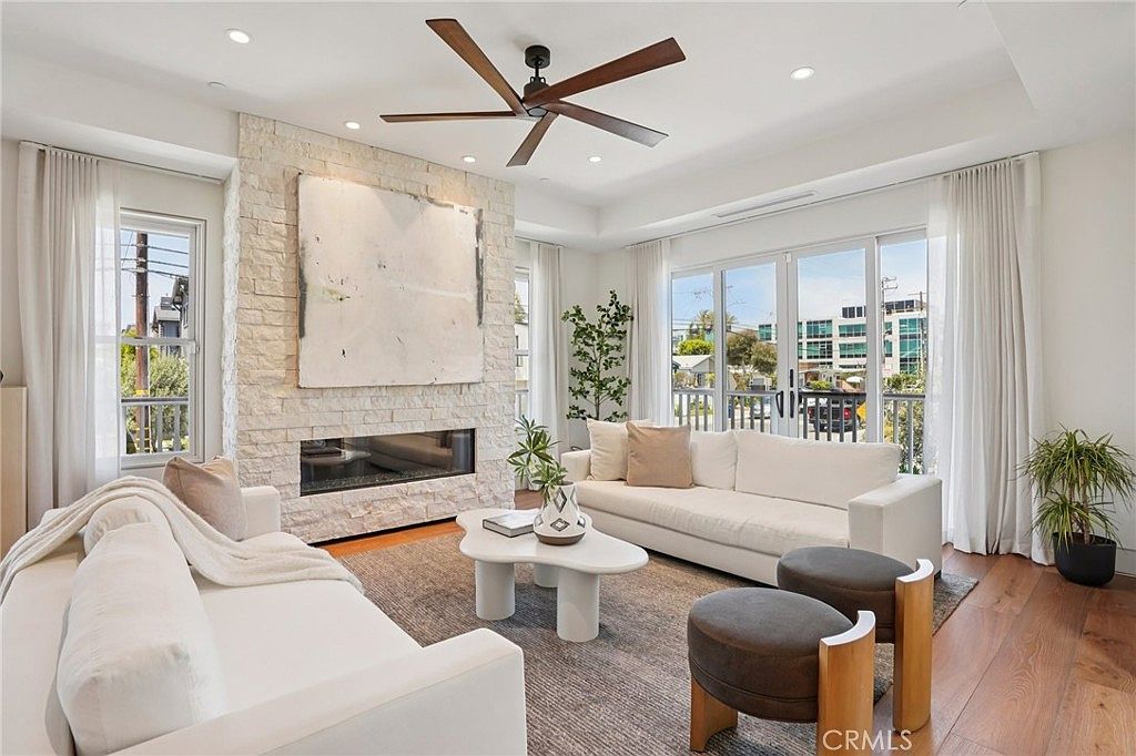 This bright and airy living room features a prominent stone-clad chimney breast that serves as a focal point, complemented by a modern linear fireplace. The space is furnished with two white sofas, a sculptural coffee table, and two dark circular ottomans, all arranged on a textured area rug over light wood flooring. Large glass doors provide ample natural light and access to an outdoor balcony, creating a seamless transition between indoor and outdoor living.