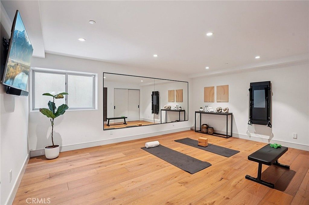 This bright and spacious home gym features light wood flooring and a large wall-mounted mirror that enhances the sense of space. The room is equipped with two yoga mats, a workout bench, a wall-mounted fitness system, and a potted plant, creating a clean and functional workout environment. A television is mounted on the wall, and recessed lighting provides even illumination throughout the area.