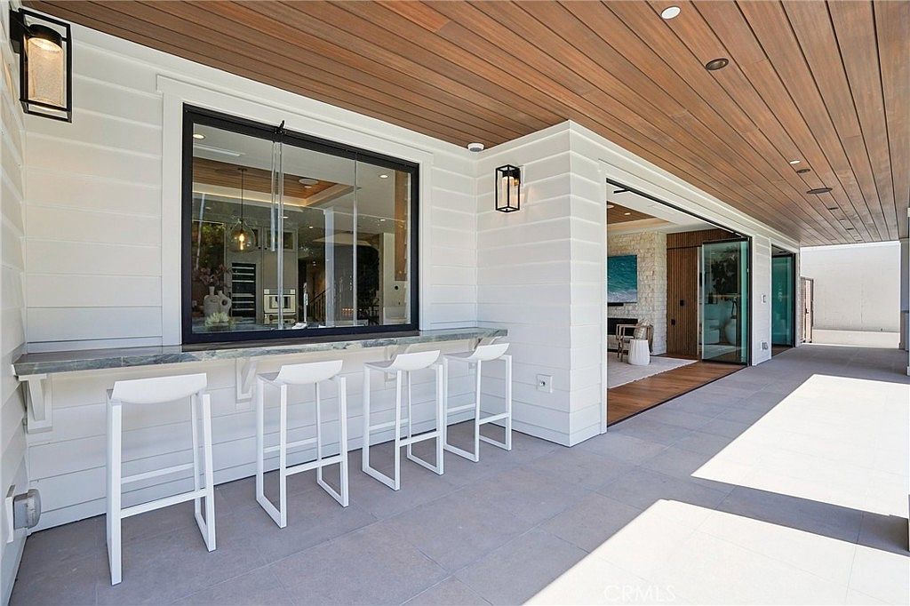 This modern outdoor patio features a covered seating area with a built-in bar counter and four white stools, perfect for entertaining. The space is defined by clean white horizontal siding, a warm wood-paneled ceiling, and large sliding glass doors that open into the interior living area. The perspective captures a seamless transition between indoor and outdoor living, highlighting the contemporary design and open-concept flow.