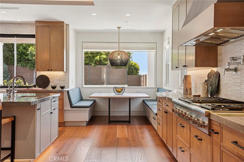 This bright and modern kitchen features a central island with a sink, light wood cabinetry, and a dedicated breakfast nook with built-in bench seating. A large window above the dining area provides ample natural light, while a professional-style range with a prominent hood serves as a focal point on the right. The space is finished with warm wood flooring and neutral-toned countertops, creating a cohesive and inviting atmosphere.