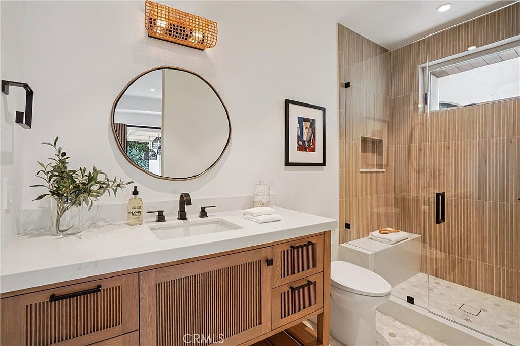 This modern bathroom features a light wood vanity with slatted cabinet doors, topped with a white countertop and a sleek black faucet. A round mirror hangs above the vanity, illuminated by a textured, cylindrical light fixture, while a glass-enclosed shower with vertical tile walls and a built-in bench sits to the right. The space is clean and contemporary, with a neutral color palette and a minimalist aesthetic.