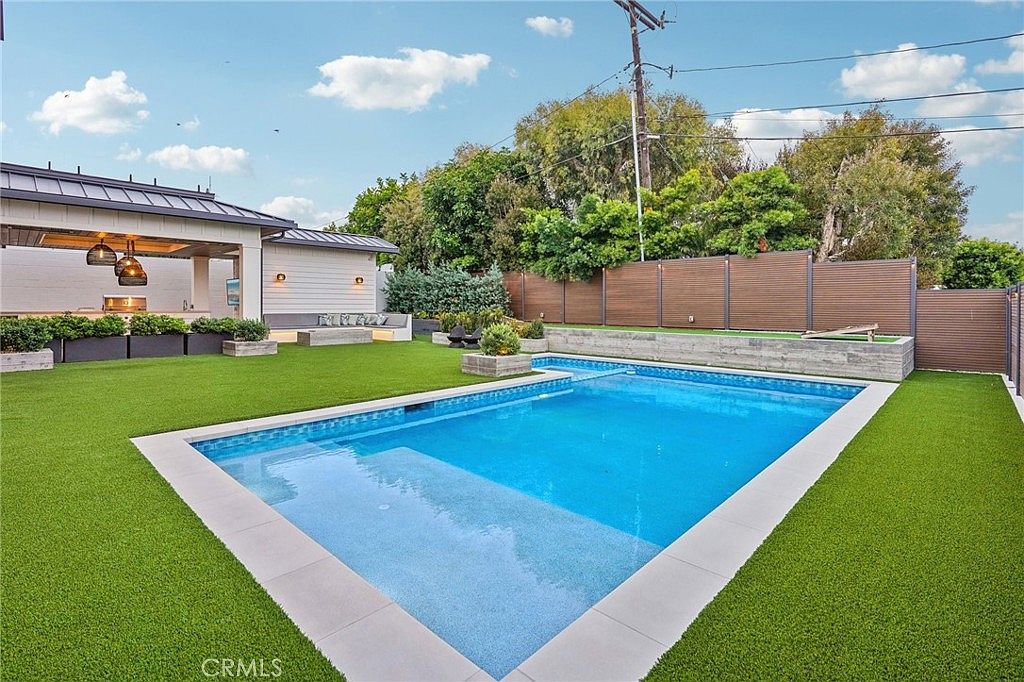 This backyard features a rectangular swimming pool with a shallow tanning ledge, surrounded by vibrant green artificial turf. A covered outdoor living area with pendant lighting and a built-in kitchen setup sits to the left, while a horizontal wood-slat fence provides privacy along the perimeter. The space is designed for modern outdoor entertaining, offering a clean, manicured aesthetic.