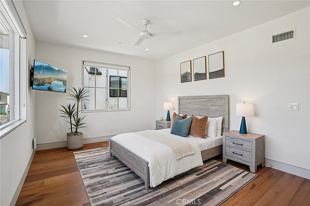 This bright and airy bedroom features light wood flooring and a neutral color palette, centered around a bed with a light-toned headboard. The room is accented by a potted plant, a wall-mounted television, and three framed art pieces above the bed, creating a clean and contemporary aesthetic. Large windows allow for ample natural light, while matching bedside tables and lamps provide a balanced, symmetrical look.