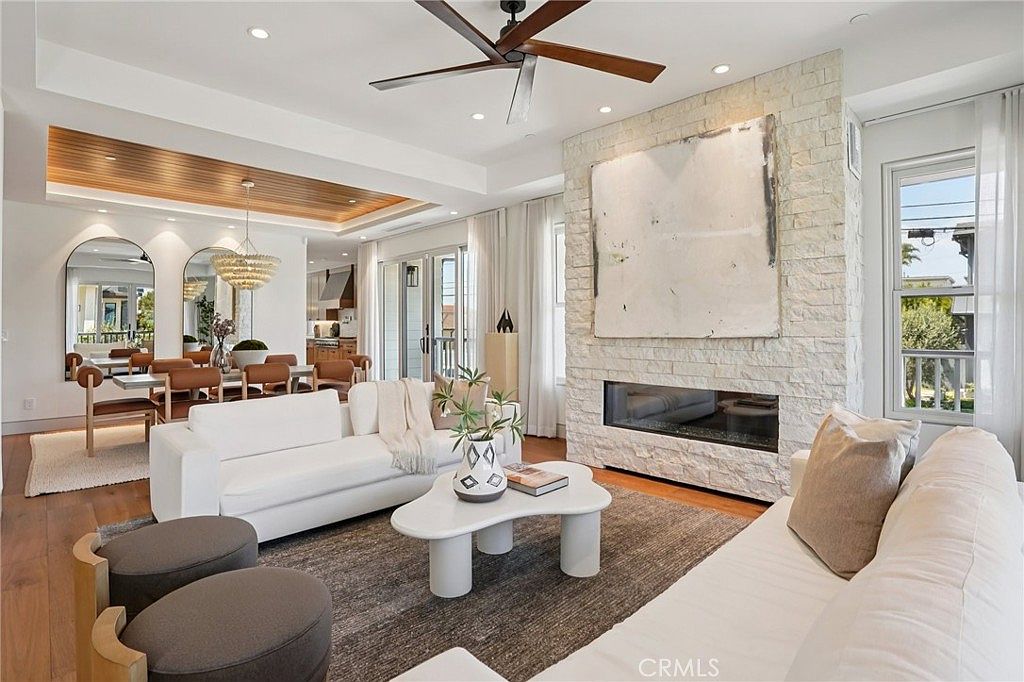 This bright and airy living room features a prominent stone-clad chimney breast that serves as a focal point, complemented by a modern linear fireplace. The space is furnished with two white sofas arranged around a sculptural, organic-shaped coffee table on a textured area rug. In the background, a dining area with a wood-paneled ceiling and a decorative chandelier creates a cohesive, open-concept flow, while large windows and glass doors invite natural light into the room.