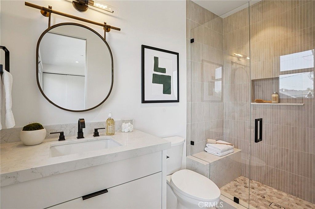 This modern bathroom features a sleek vanity with a stone countertop, a round mirror, and matte black fixtures. To the right, a glass-enclosed shower showcases vertical wall tiles, a built-in stone bench, and a pebble-style floor. The space is bright and minimalist, offering a clean, contemporary aesthetic.