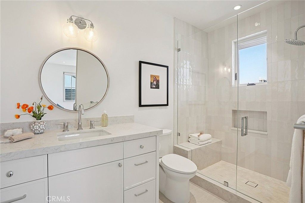 This bright and modern bathroom features a white vanity with a stone countertop, a circular mirror, and a glass-enclosed walk-in shower with light-colored wall tiles. A built-in bench is integrated into the shower area, which also includes a recessed niche for storage. The space is well-lit by a dual-bulb vanity light and a window above the shower, creating a clean and airy atmosphere.