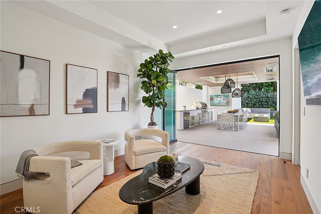 This bright and airy living room features a contemporary design with light wood flooring and two cream-colored armchairs arranged around a dark, organic-shaped coffee table. A large, open-concept transition leads to a covered outdoor patio, creating a seamless indoor-outdoor living experience. The space is decorated with minimalist wall art and a tall indoor plant, emphasizing a clean, sophisticated aesthetic.