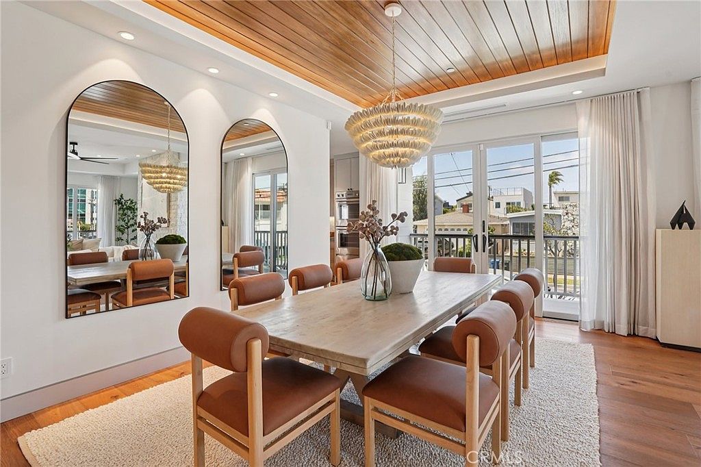 This elegant dining room features a light wood table surrounded by six modern chairs with tan leather-style upholstery. The space is defined by a striking wood-paneled ceiling, a large textured chandelier, and two arched mirrors that reflect the room's bright, airy atmosphere. Large glass doors lead to an outdoor balcony, allowing natural light to fill the room, which is anchored by a plush, light-colored area rug over hardwood floors.
