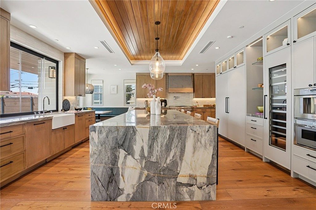 This spacious, modern kitchen features a large central island with a dramatic, veined stone waterfall edge and light wood cabinetry. The room is anchored by a warm wood-paneled ceiling inset with pendant lighting, creating a sophisticated and inviting atmosphere. Large windows provide ample natural light, while high-end appliances and a farmhouse-style sink complete this high-end culinary space.