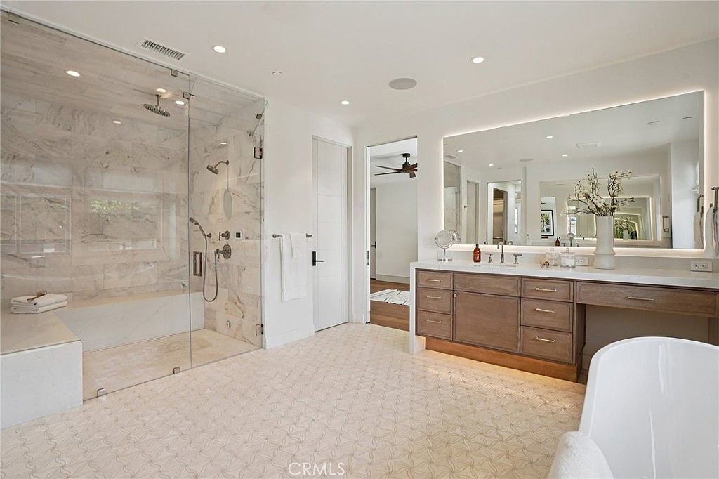 This spacious primary bathroom features a large walk-in shower with glass walls and light-toned stone tile, alongside a double vanity with a wide mirror and integrated lighting. The room is finished with patterned tile flooring and a freestanding tub visible in the foreground, creating a bright and luxurious atmosphere. The perspective is wide-angle, capturing the entire layout from the entrance toward the vanity and shower area.