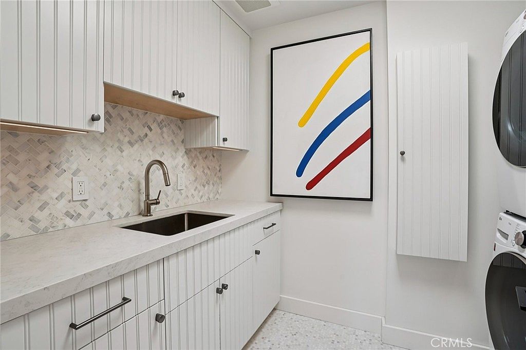 This bright and modern laundry room features white vertical-paneled cabinetry and a light-toned stone countertop with an integrated sink. A patterned tile backsplash adds visual interest, while a large, colorful abstract art piece serves as a focal point on the wall. The space is equipped with a stacked washer and dryer unit and includes a tall, narrow storage cabinet, creating a clean and organized utility area.