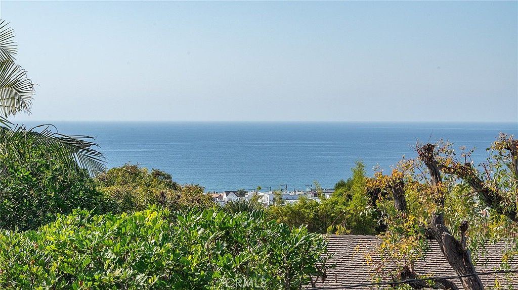This elevated outdoor perspective captures a sweeping, panoramic view of the ocean horizon framed by lush green foliage and trees in the foreground. Below the tree line, a glimpse of coastal residential rooftops is visible, suggesting a prime hillside location. The scene conveys a serene and tranquil atmosphere, emphasizing the property's scenic coastal vantage point.