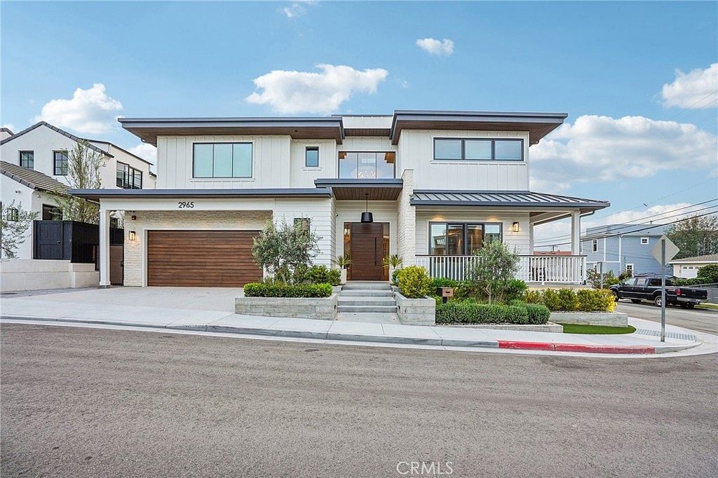 This modern two-story residence features a striking contemporary design with a mix of horizontal wood siding, white vertical paneling, and light-colored stone accents. The front facade includes a large wooden garage door, a prominent central entryway with a modern door, and a covered porch area with a white railing. The property is situated on a corner lot with manicured landscaping, concrete steps leading to the entrance, and a clear view of the surrounding neighborhood under a bright blue sky.
