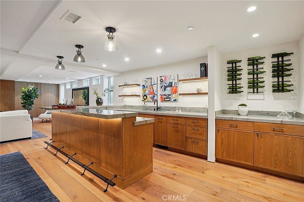 This modern kitchen features a large island with a stone countertop and integrated lighting, providing ample space for seating and food preparation. The space is accented by warm wood cabinetry, a wall-mounted wine rack, and contemporary pendant lighting. The open-concept design flows seamlessly into the adjacent living area, creating a bright and inviting atmosphere.