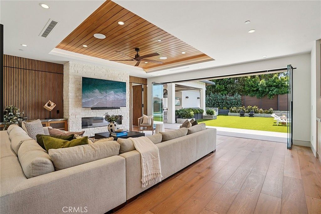 This spacious, open-concept living room features a warm wood-paneled ceiling with a central fan and a prominent stone-clad chimney breast housing a television and fireplace. A large, neutral-toned sectional sofa anchors the room, which seamlessly transitions to an outdoor patio through expansive, retractable glass walls. The light wood flooring and clean, modern aesthetic create an inviting atmosphere that blends indoor comfort with the adjacent landscaped yard.