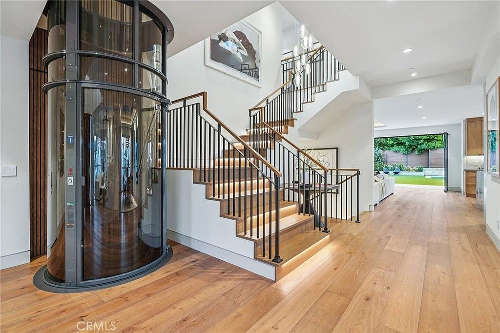 This modern entryway features a striking cylindrical glass elevator as a central architectural focal point, positioned next to a staircase with light wood treads and black metal railings. The open-concept design leads the eye toward a bright living area with large glass doors that open to a backyard, creating a seamless transition between indoor and outdoor spaces. The light wood flooring and neutral color palette contribute to a clean, sophisticated, and airy atmosphere.