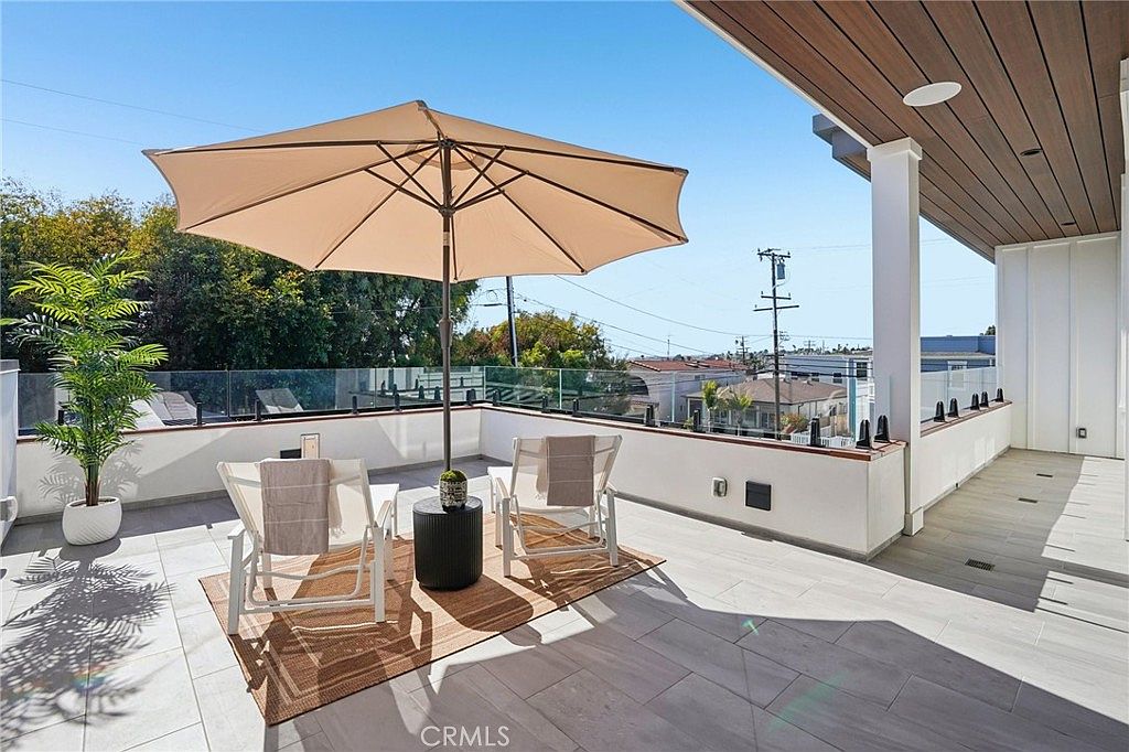 This modern rooftop balcony features light-toned tile flooring and a glass railing that provides an unobstructed view of the surrounding neighborhood. The space is furnished with two white lounge chairs, a small side table, and a large beige umbrella, creating a comfortable outdoor seating area. A covered section with a wood-paneled ceiling offers shade, while a potted plant adds a touch of greenery to the minimalist design.