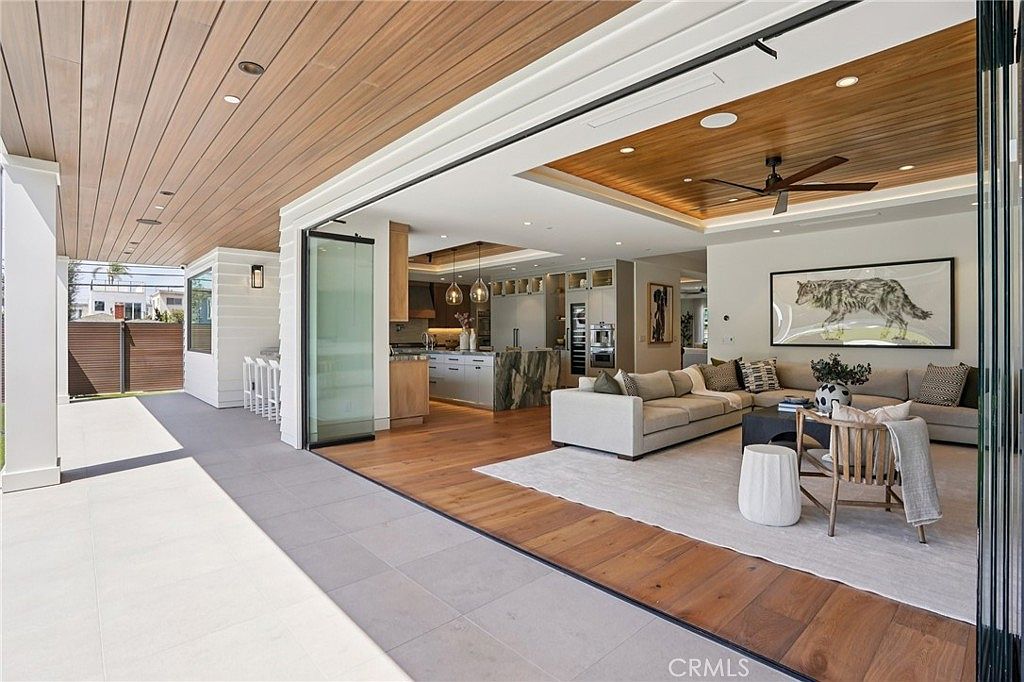 This open-concept living space seamlessly transitions to an outdoor patio through expansive, retractable glass walls, creating an indoor-outdoor living experience. The interior features a coffered wood-paneled ceiling with recessed lighting, a large sectional sofa, and a neutral area rug, while the adjacent kitchen showcases a stone-topped island and modern cabinetry. The perspective captures the fluid connection between the refined indoor living area and the covered outdoor lounge space.