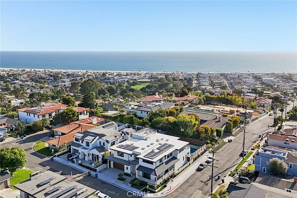 This high-angle aerial view captures a contemporary multi-level residence situated in a coastal neighborhood. The property features a flat roof with integrated solar panels, a private swimming pool, and a multi-car garage, all set against a backdrop of the Pacific Ocean. The surrounding area is densely populated with residential homes, lush greenery, and paved streets, emphasizing the prime location near the beach.