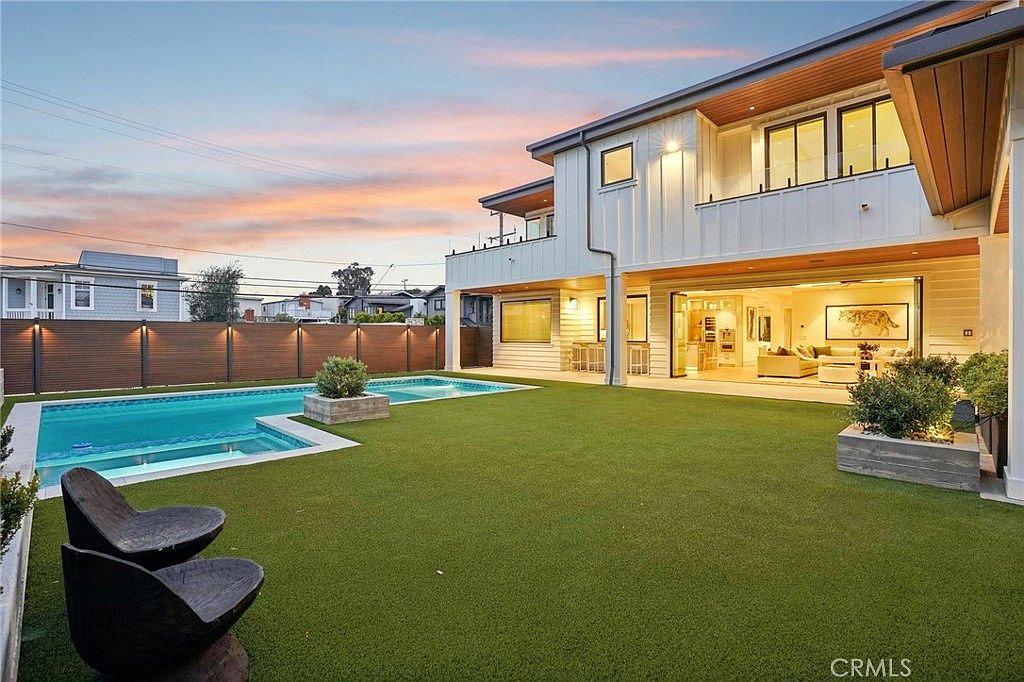 This rear view of a modern two-story home showcases a seamless transition between indoor and outdoor living spaces through large, open glass doors. The backyard features a pristine swimming pool with an integrated spa, a manicured artificial turf lawn, and contemporary landscaping. The exterior is finished in white vertical siding, complemented by warm wood-toned accents and ambient lighting that creates an inviting atmosphere at dusk.