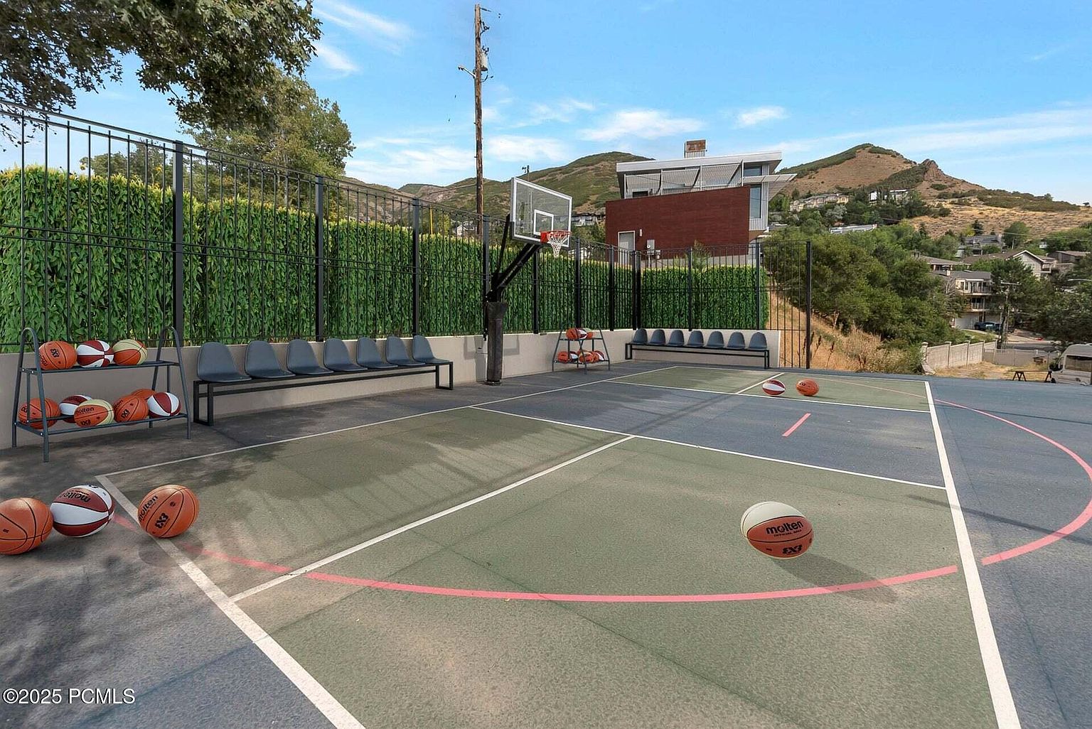 This image showcases a well-maintained outdoor basketball court, complete with multiple basketballs, seating, and a sturdy fence. The court features a combination of green and blue surfaces, with clearly marked lines and a hoop. The backdrop includes lush greenery and a view of the surrounding hills, adding to the appeal of this community amenity.