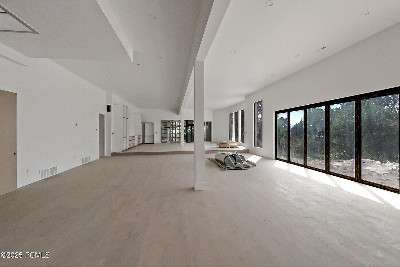 This is a spacious, modern living room with light wood flooring and white walls, creating a bright and airy atmosphere. Large sliding glass doors offer expansive views of the surrounding landscape, while minimalist architectural details and recessed lighting contribute to the clean aesthetic. The room is currently unfurnished, highlighting its potential and size.