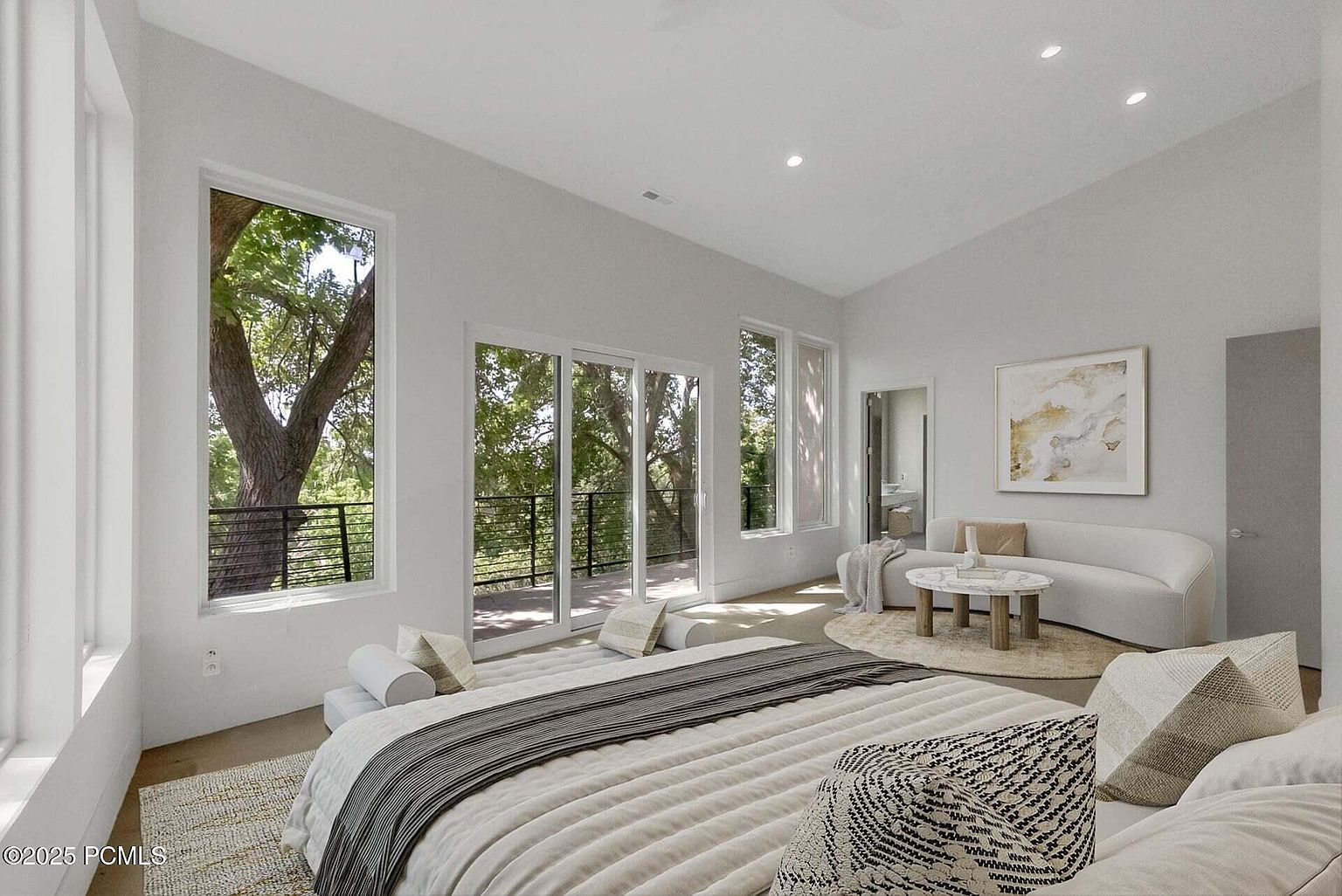 This is a bright and airy primary bedroom featuring a large bed with decorative pillows and a gray throw. The room has large windows and sliding glass doors that lead to a balcony, offering views of the surrounding trees. A curved sofa and round coffee table create a cozy seating area, complemented by a neutral color palette and modern artwork.