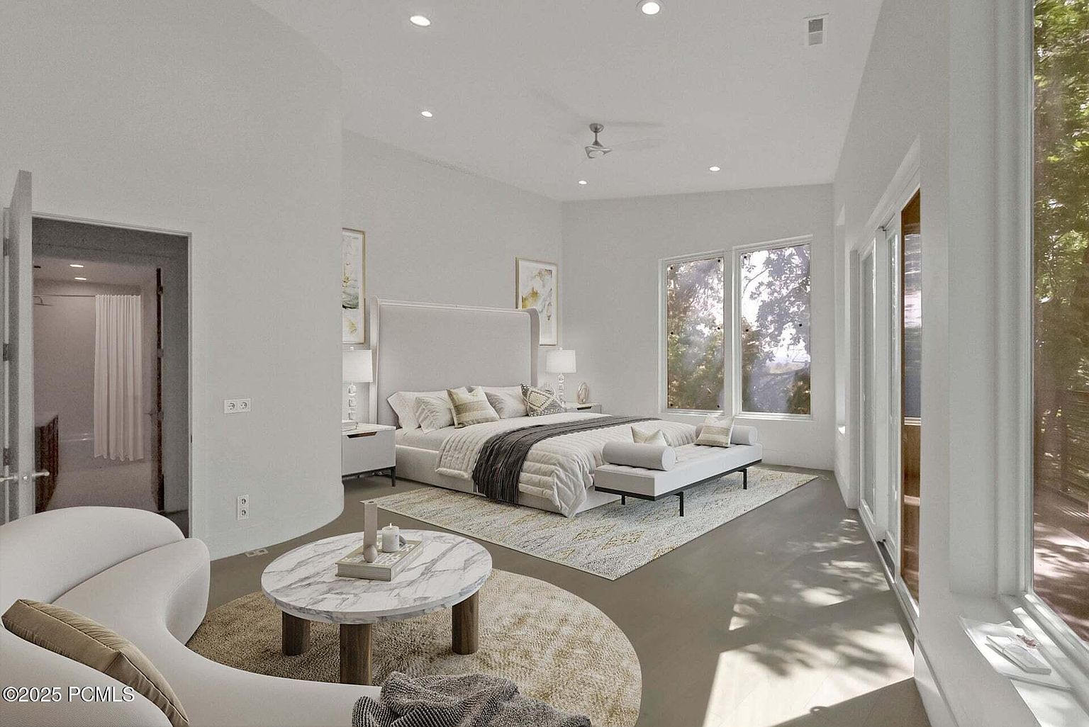 This is a spacious primary bedroom featuring a large bed with a high headboard, neutral-toned bedding, and a seating area with a round marble-topped table. The room is well-lit with natural light from large windows and recessed lighting, creating a bright and airy atmosphere. The style is modern and luxurious, with a focus on comfort and relaxation.