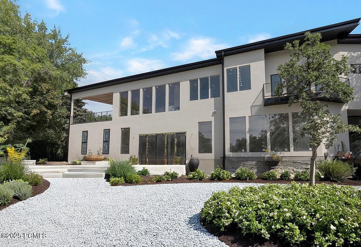 This is a rear view of a modern two-story home with a light-colored exterior. The house features large windows and doors, a balcony, and a well-maintained yard with white gravel and various plants. The overall impression is one of contemporary elegance and outdoor living.