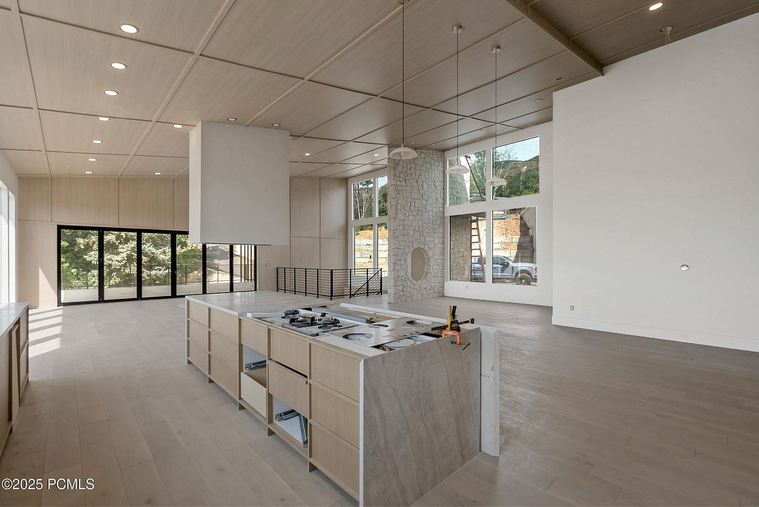 This is a spacious, modern kitchen featuring light wood cabinetry and a large central island with a cooktop. The room is well-lit with natural light from large windows and recessed lighting, and a unique stone fireplace adds a touch of rustic charm. The open floor plan and clean lines create a contemporary and inviting atmosphere.