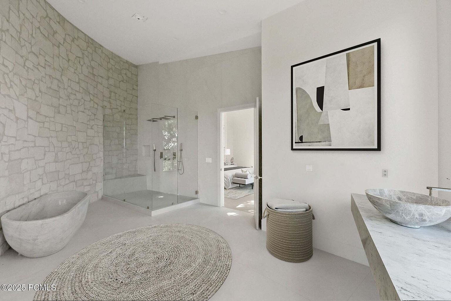 This is a luxurious primary bathroom featuring a stone accent wall, a freestanding soaking tub, and a glass-enclosed shower. The room is decorated with a neutral color palette, a round woven rug, and a modern art piece, creating a spa-like atmosphere. A marble countertop with a vessel sink adds to the high-end feel of the space.