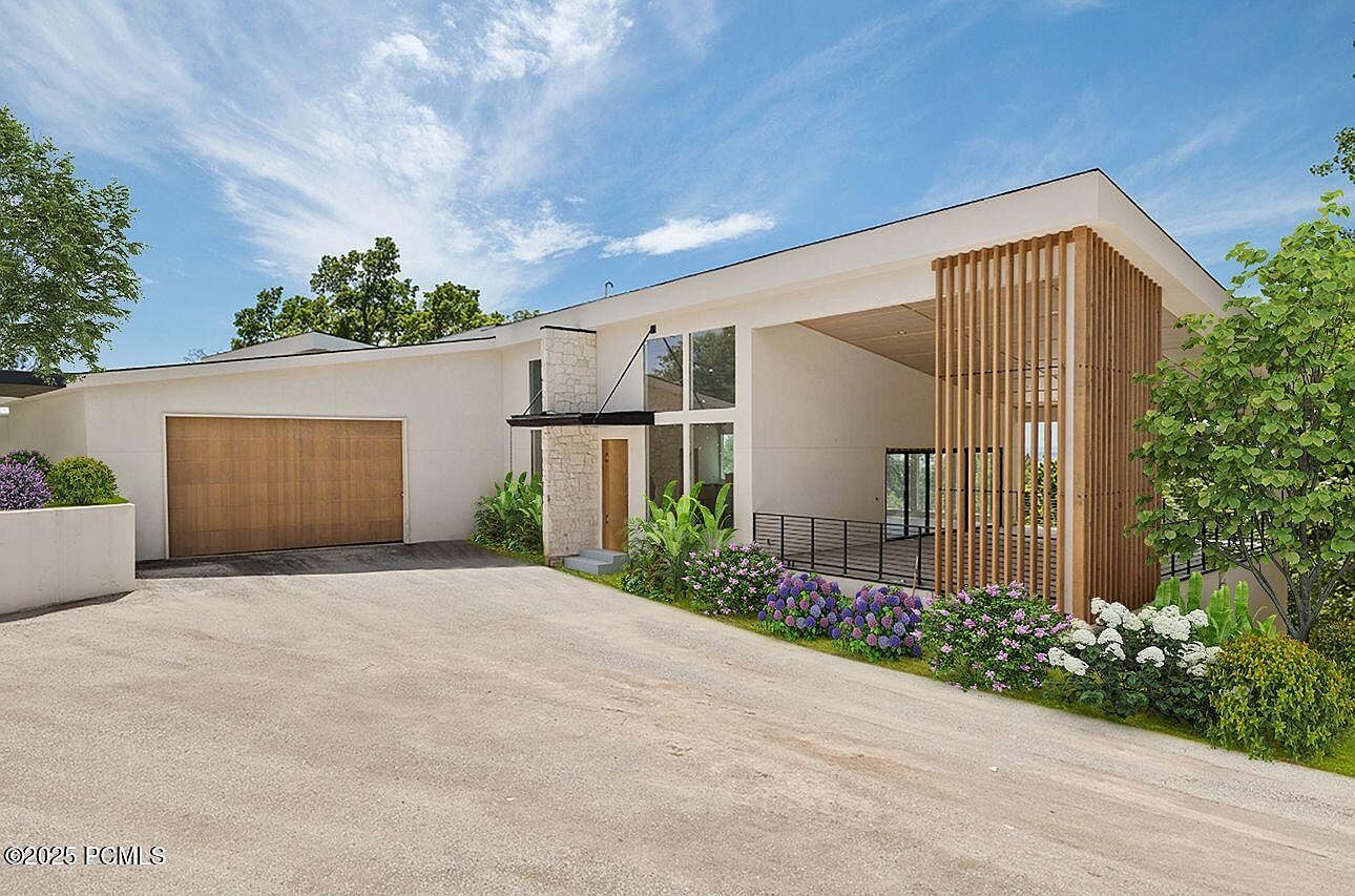 This is a front exterior view of a modern, single-story home. The house features a light-colored facade, a wooden garage door, and a unique wooden slat design element near the entrance. Landscaping includes flowering plants and greenery, adding to the property's curb appeal.