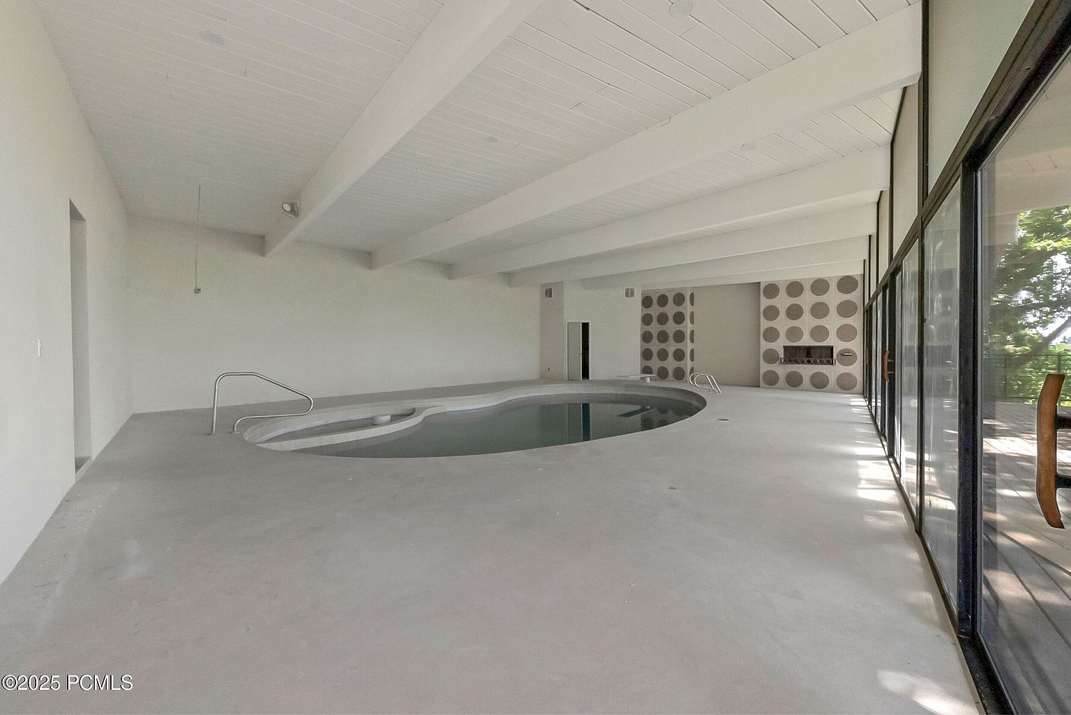 This interior shot showcases a unique indoor pool area. The pool is kidney-shaped and surrounded by a light gray concrete floor. The room features a white beamed ceiling, a decorative wall with circular patterns, and large windows that provide natural light and a view of the outdoors. The overall impression is modern and spacious.