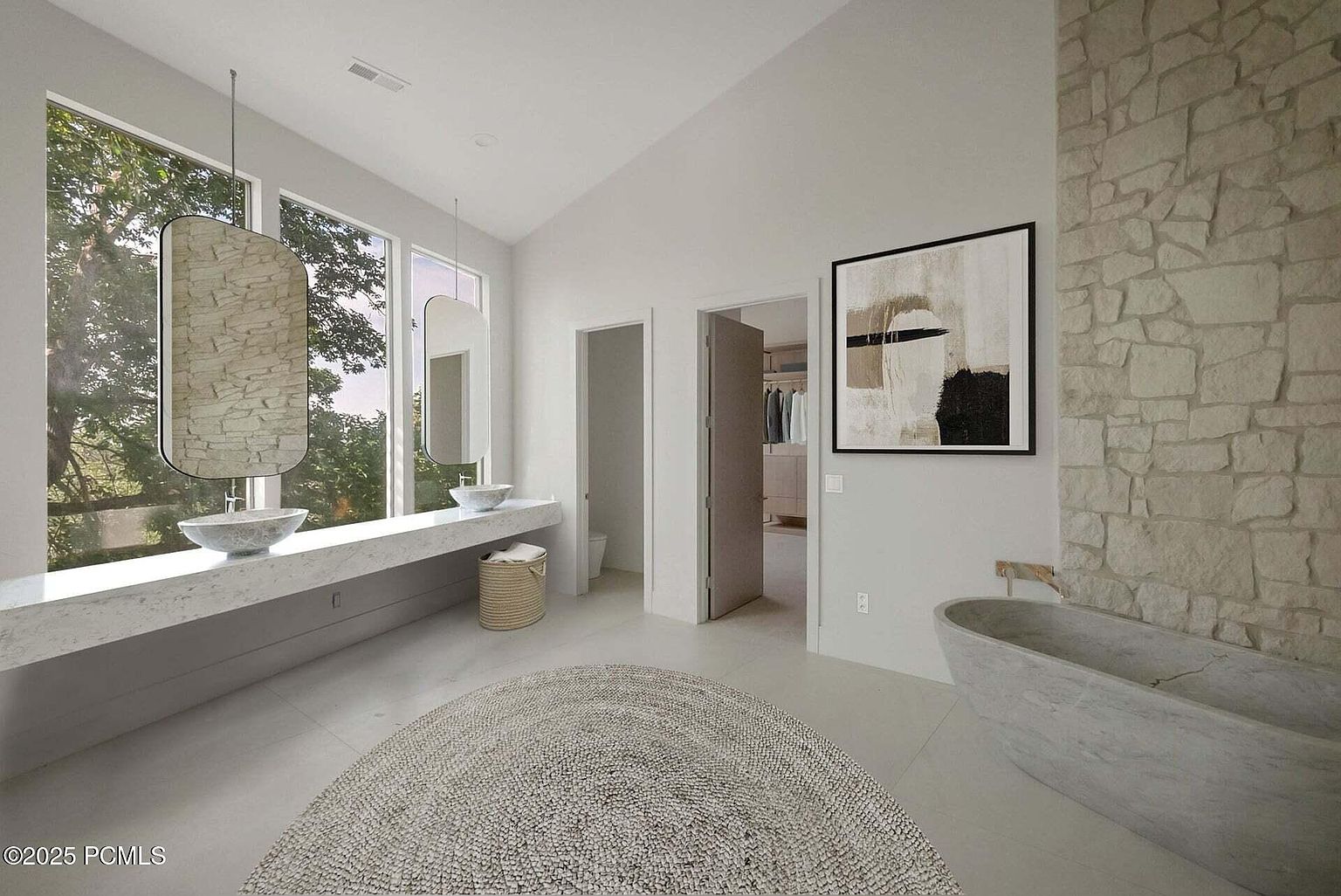 This is a bright and modern primary bathroom featuring dual vessel sinks on a long, light-colored countertop, complemented by unique hanging mirrors. A freestanding stone bathtub sits against a textured stone wall, adding a spa-like ambiance. The room is spacious with a neutral color palette and a round woven rug adding texture to the tile floor.