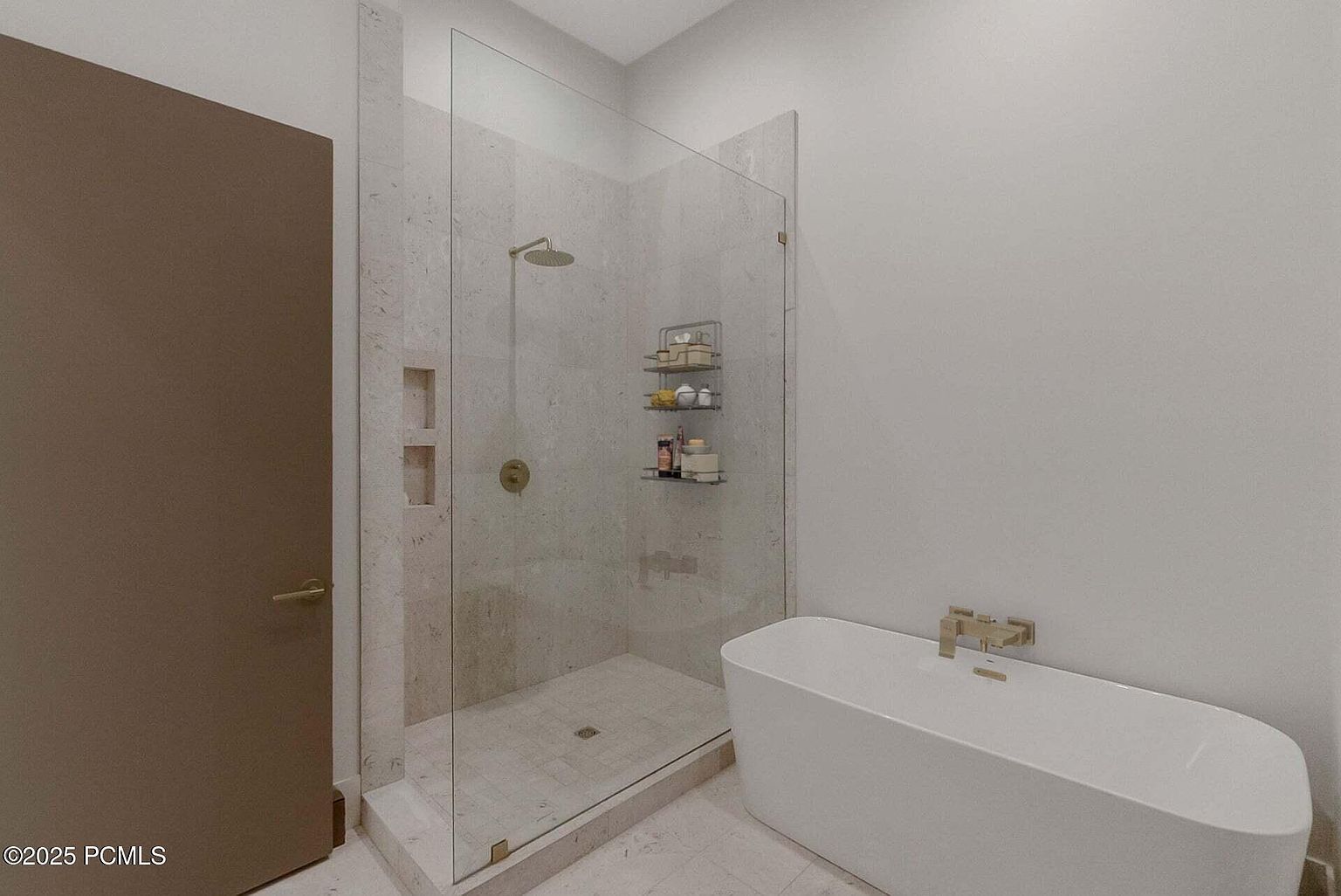 This is a modern primary bathroom featuring a glass-enclosed shower with a rainfall showerhead and built-in shelving. A freestanding white bathtub with gold fixtures sits adjacent to the shower. The bathroom has a clean and minimalist design with light-colored tile flooring and walls, creating a spa-like atmosphere.