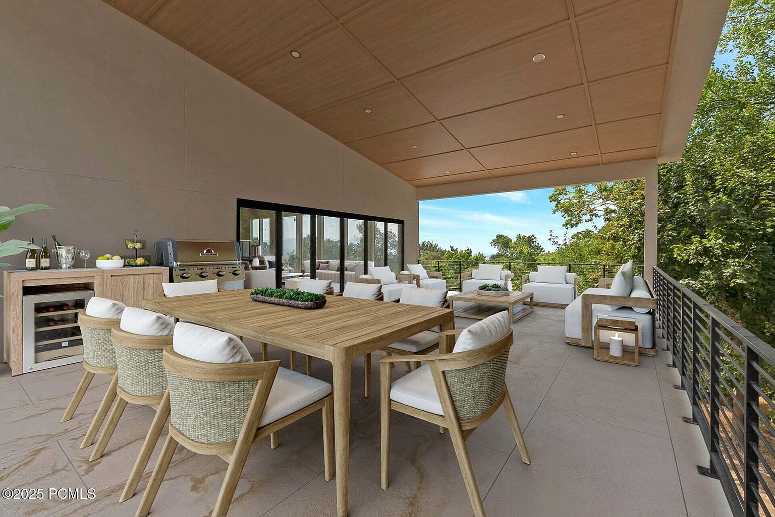 This image showcases a spacious outdoor patio or balcony area, furnished with a large wooden dining table and chairs, as well as comfortable lounge seating. The space features a built-in bar area with a wine fridge and a grill, suggesting an ideal setting for outdoor dining and relaxation. The open design offers views of the surrounding greenery and sky, enhancing the appeal of this outdoor living space.