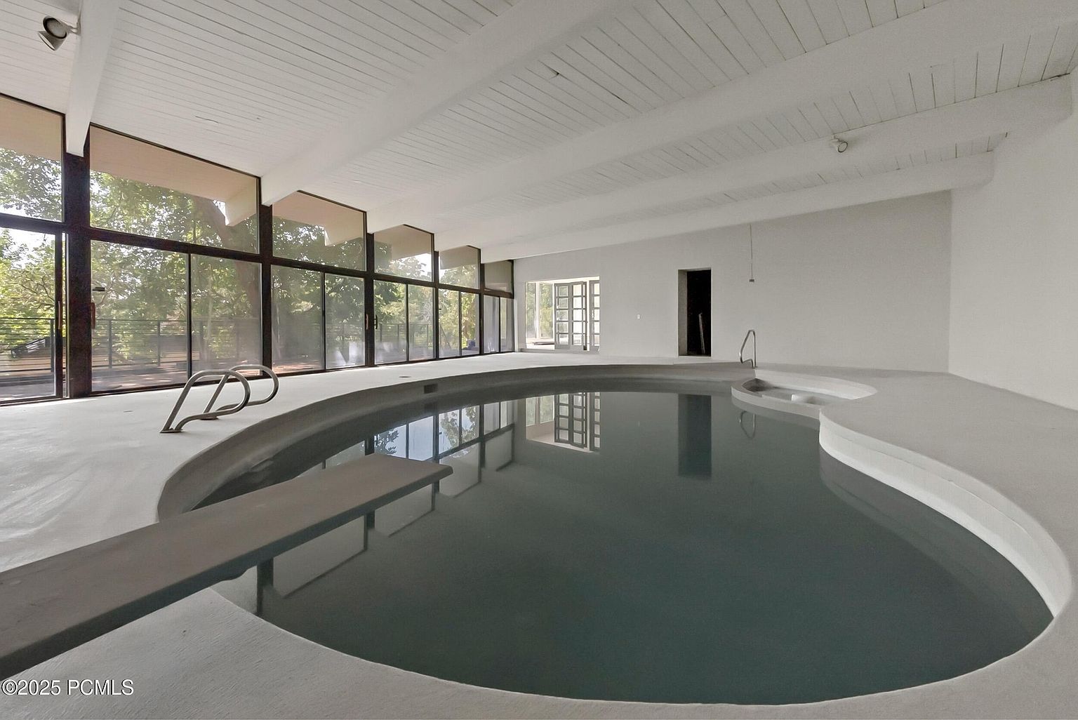 This image showcases an indoor pool area, featuring a large, uniquely shaped pool with a diving board and integrated steps. The space is bright and airy, thanks to the expansive windows that line one side, offering views of the outdoors. The white walls and ceiling create a clean, modern aesthetic, enhancing the sense of spaciousness and relaxation.