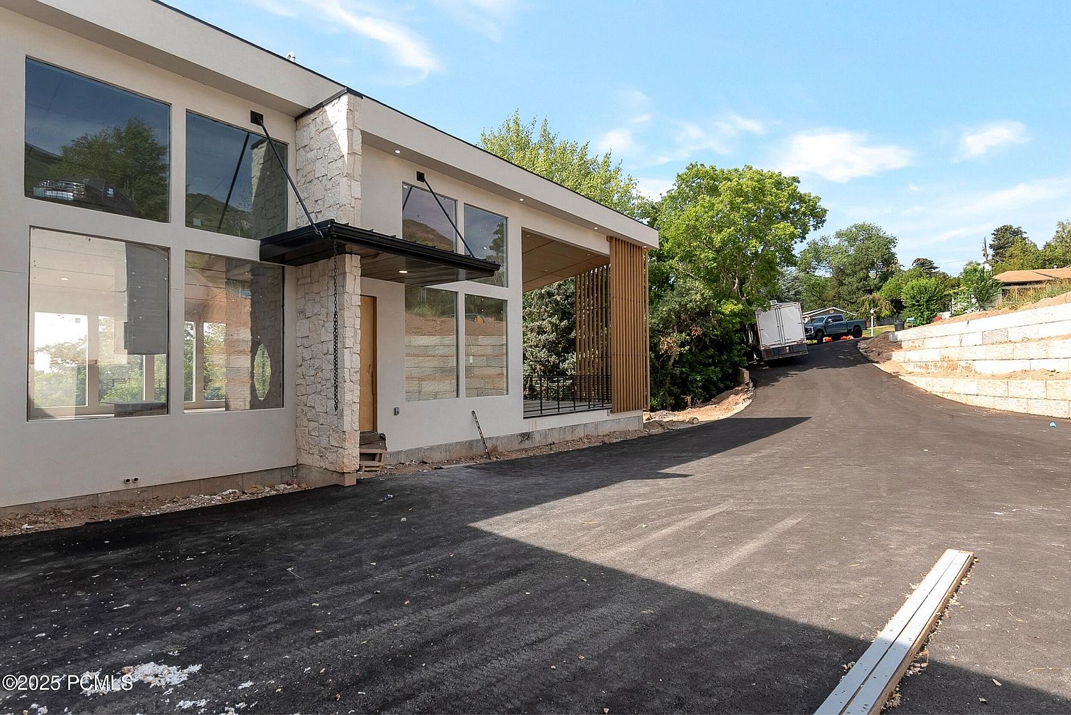 This is a front exterior view of a modern home with large windows and a stone accent wall. The house features a flat roof and a covered entryway. A newly paved driveway leads up to the house, and there is some landscaping visible in the background. The overall impression is clean, contemporary, and well-maintained.