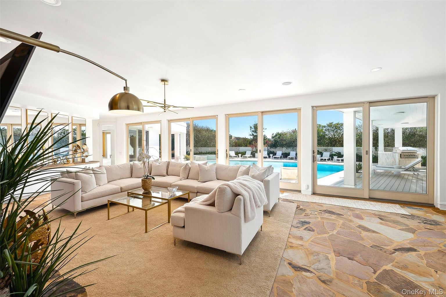 This is a bright and spacious living room featuring a large sectional sofa, glass coffee tables, and floor-to-ceiling windows that offer a view of the pool area. The room is decorated in neutral tones with a mix of carpet and stone flooring, creating a luxurious and inviting atmosphere. The open layout seamlessly connects the indoor and outdoor spaces.
