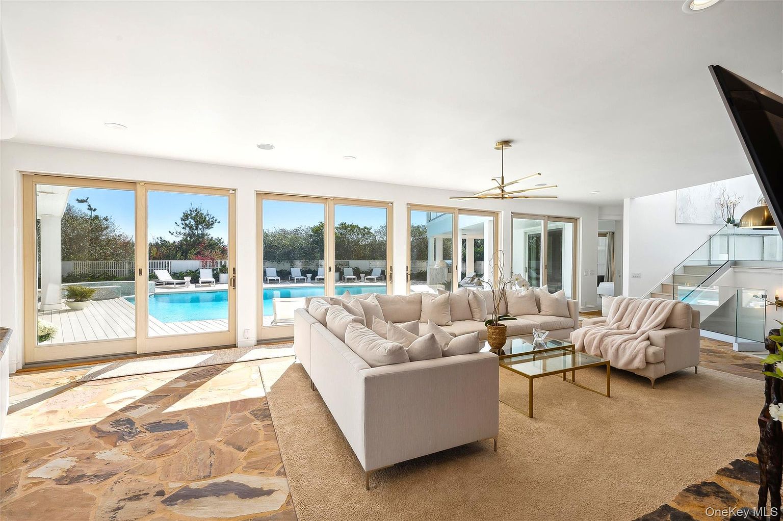 This is a bright and spacious living room featuring a large sectional sofa, a glass coffee table, and a beige area rug. The room has large sliding glass doors that offer views of the pool area, bringing in natural light. A modern chandelier hangs from the ceiling, and a staircase is visible in the background, adding to the luxurious feel of the home.
