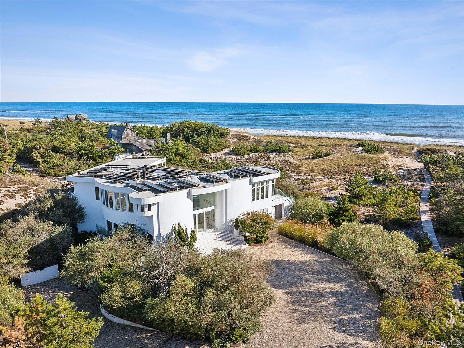 This aerial view showcases a modern, white beachfront home with a curved design and solar panels on the roof. The property is surrounded by lush greenery and a sandy beach leading to the ocean. A winding driveway provides access to the house, enhancing its secluded and luxurious appeal.