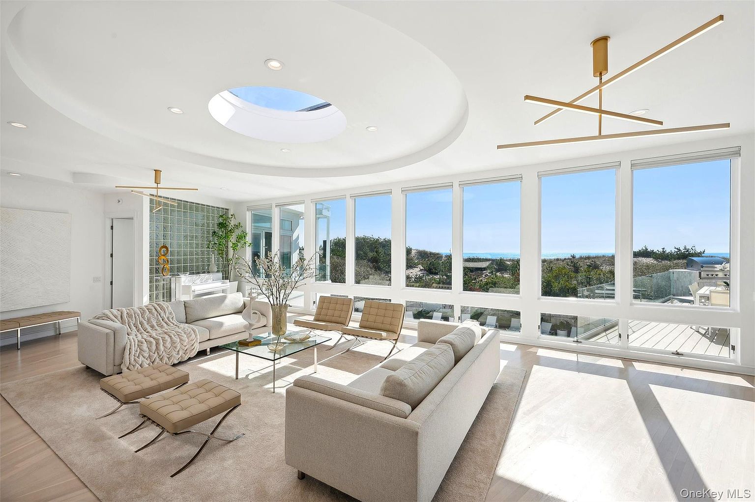 This is a bright and airy living room featuring large windows with ocean views. The room is furnished with modern sofas, chairs, and a glass coffee table, all set on a neutral-toned rug. A unique circular skylight and modern chandelier add architectural interest, creating a luxurious and inviting space.