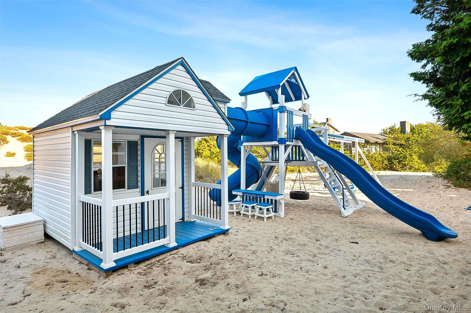 This image showcases a charming backyard play area, featuring a white and blue playhouse with a small porch and a separate playset with a slide, swing, and climbing structure. The area is covered in sand, creating a beach-like atmosphere, and is surrounded by lush greenery, suggesting a private and family-friendly outdoor space. The scene is captured from a medium-distance perspective, highlighting the play area's integration within the yard.