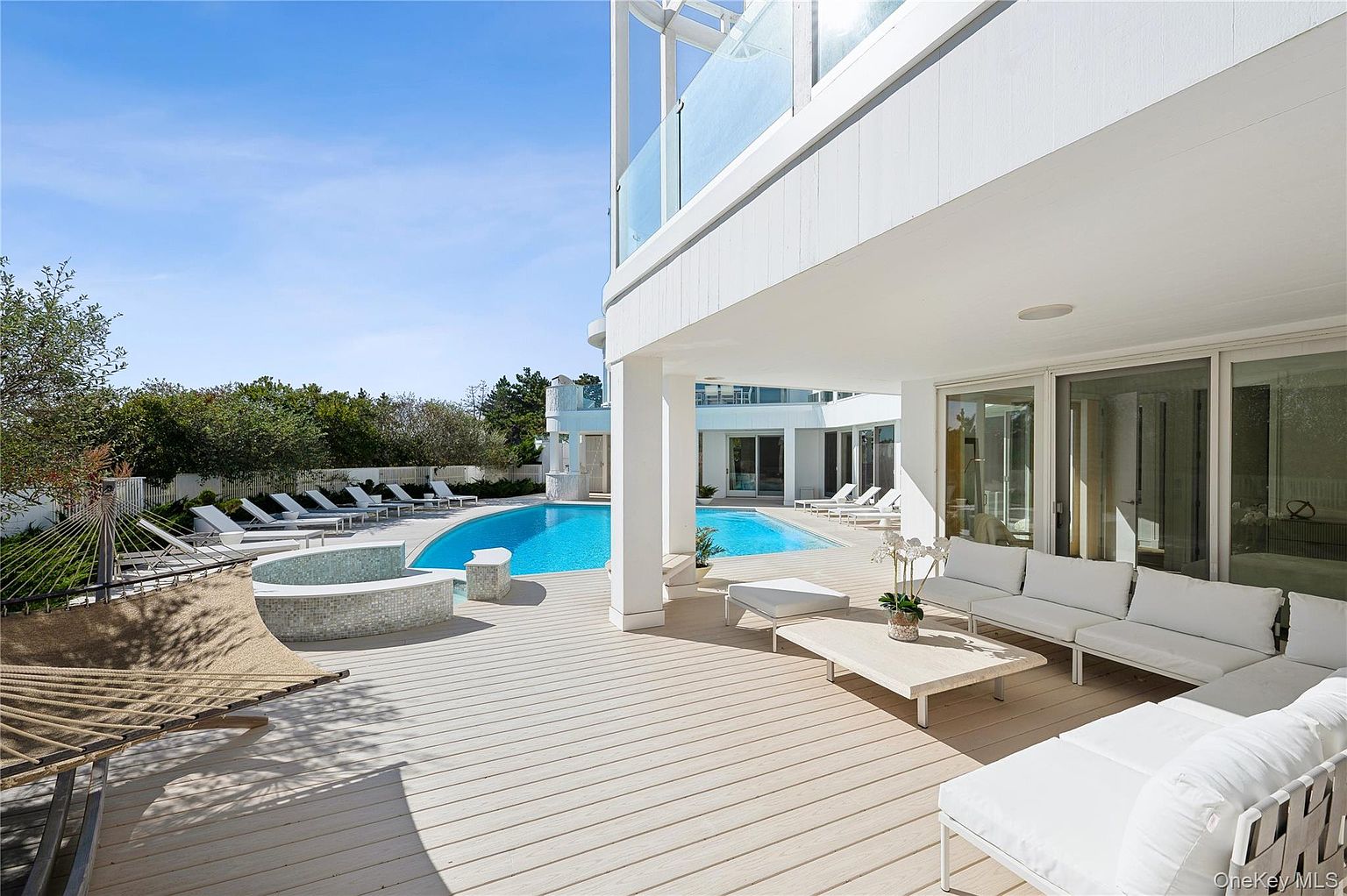 This image showcases a luxurious outdoor living space with a modern aesthetic. The patio features sleek white furniture, a light-colored wooden deck, and a view of a stunning pool and spa area. The architectural design of the house is contemporary, with clean lines and a bright, airy feel, perfect for relaxation and entertainment.